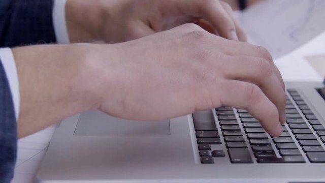 Close up of businessman hands are typing the data on laptop keyboard. Professional traider is pushing the bottons by his fingers and his female colleague, who is sitting near, checking rates on graph