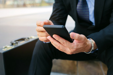 close up of young business man wearing modern black suit using mobile smart phone and briefcase sitting on walkway in city, internet network connection, technology communication and financial concept