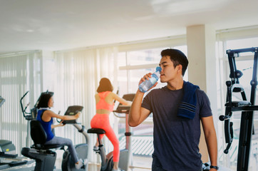 Fototapeta na wymiar muscular asian young handsome man with towel drinking water while resting after workout for good healthy in fitness gym with woman, bodybuilder, healthy lifestyle, exercise and sport training concept