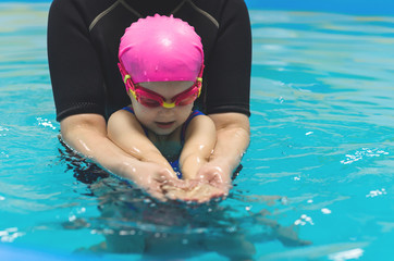 A little girl of European appearance in a pink rubber cap learning to swim in the pool