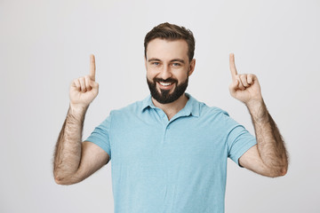 Portrait of happy bearded caucasian guy pointing up with both hands and smiling cheerfully, standing over gray background. Man shows city banner with his photo on it to his friends