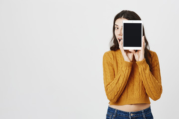 Indoor shot of excited and happy woman hiding face behind tablet as if promoting it, peeking and expressing amazement over gray background. Girl finally found great present for mother