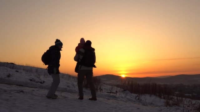 A happy family, mom, father and daughter play on the mountain in the winter in the rays of the setting sun. A man is holding a child in his arms.