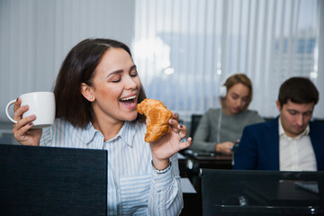 Relaxed female student with mug of tea or coffee and snack croissant and apple. Group of people on background. Happy woman having lunch on working place. People, business, education concept