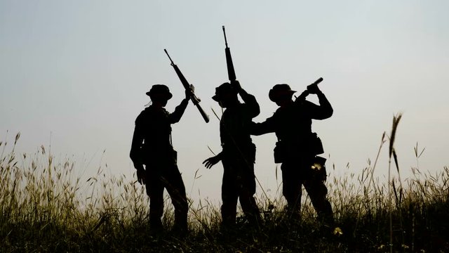 Silhouette of three fully equipped and armed soldiers standing in battle field after the war ends.