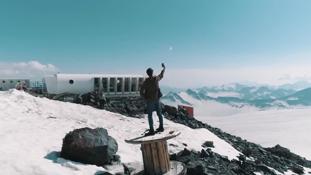 Aerial shot young stylish man in sunglasses makes selfie standing on wooden cable spool on rocks at snowy mountains picturesque landscape. Sunny day, blue sky