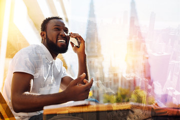Emotional talk. Cheerful emotional attentive student feeling happy and laughing while sitting alone at the table in a lovely cafe and talking on the phone with his parents