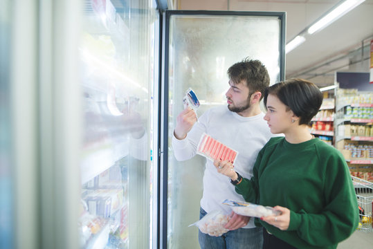 A beautiful young couple chooses frozen foods near the refrigerator in a supermarket. A couple looks at the labels of the goods in the store.