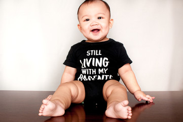 A baby boy is wearing a shirt with a message saying he is still living with my parents