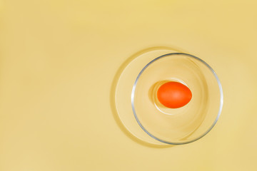 One Easter orange egg is lying in a transparent glass cup on a yellow background with copy space.