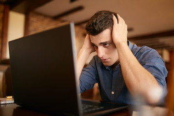 Feeling tired. Frustrated young handsome man looking exhausted and sick while sitting at his working place with laptop on the table. Freelancer has headache and holds his head both hands.