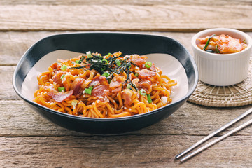 Korean hot and spicy instant noodle in black and white bowl topped with bacon, sesame, seaweed and chopped scallion served with kimchi on wood table in side view. Homemade delicious asian food concept