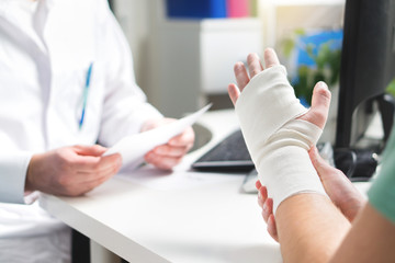 Injured patient showing doctor broken wrist and arm with bandage in hospital office or emergency room. Sprain, stress fracture or repetitive strain injury in hand. Nurse helping customer. First aid. - obrazy, fototapety, plakaty