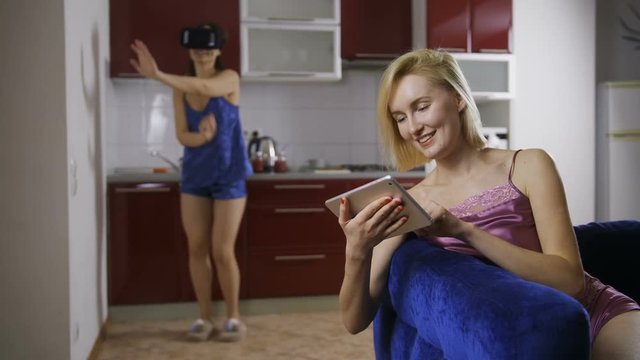 Young brown hair woman in lingerie using virtual reality glasses and dancing at home in the kitchen while her blonde friend sitting on the sofa, using digital tablet and looking at her. Dolly shot