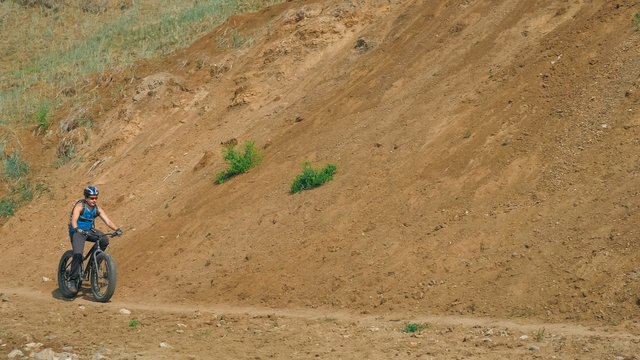 Fat bike also called fatbike or fat-tire bike in summer driving on the road. The guy rides by the hill on a sand clay path, behind him the shore by the sea. - Powered by Adobe