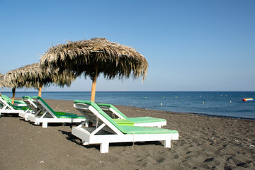 Volcanic beaches with black sand of the island of Santorini and white chaise-longue