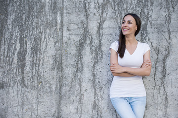 Great day. A young smiling woman in a white T-shirt is standing at a concrete wall with her hands crossed