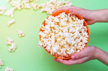 Popcorn in an orange bowl in children's hands on a green background. Top view