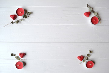 Fototapeta na wymiar Red candies in a form of heart, branch of eucalyptus and red candles on the white wooden table. Background for greeting card on Valentine's day with place for text.