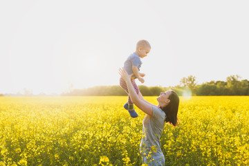 Fototapeta premium Joyful woman walk on green yellow flowering field background, rest, have fun, play, toss up little cute child baby boy. Mother, little kid son. Family day 15 of may, love, parents, children concept.