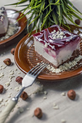 A piece of vanilla cake with chocolate base and cherry glaze decorated with some pieces of white chocolate served in a brown clay dish with silver fork placed on a beige tablecloth. Blurred background