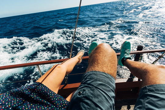 Young woman in colorful dotted dress with ballerinas and man in shorts with turquoise shoes sitting on board of a sailing boat with suntanned legs on a taffrail while sailing and watching the ocean