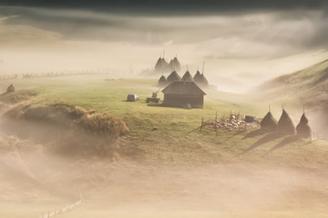 Beautiful rural mountain landscape in the morning light with fog, old houses and haystacks,...