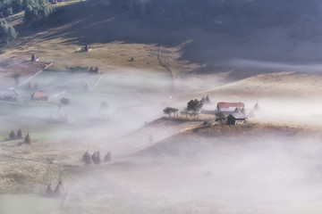 Beautiful rural mountain landscape in the morning light with fog, old houses and haystacks, Fundatura Ponorului, Hunedoara County, Romania