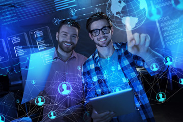 Cheerful team. Excited young clever programmers smiling while standing in front of a futuristic transparent screen with documents and gadgets in their hands