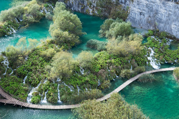 A boardwalk cuts right beside a long series of waterfalls in Plitvice Lakes National Park. The water is clear and turquoise and there are white cliffs in the background. This is an aerial shot.