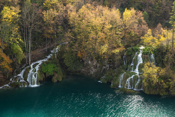 An aerial picture of two waterfall and a hiking trail in Plitvice Lakes National Park. The trees are losing leaves and changing colors in the fall. The water is a turquoise blue and the grass is lush.