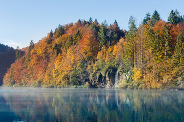 A waterfall flows into a turquoise lake in autumn. The trees are red orange green yellow and the sky is clear. There is a layer of mist or fog on top of the still water.