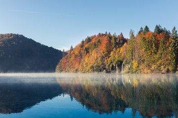 Early morning on a calm lake in Plitvice Lakes National Park. In the background there is a waterfall flowing into the lake. The trees are colored red orange green and yellow. There is a layer of fog.