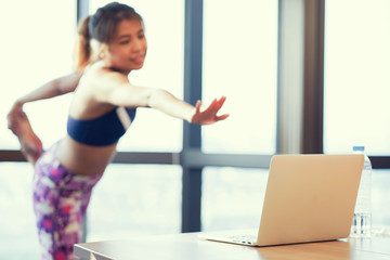 close up on notebook laptop with fitness woman stretching body before yoga workout exercise at condominium room , healthy lifestyle concept