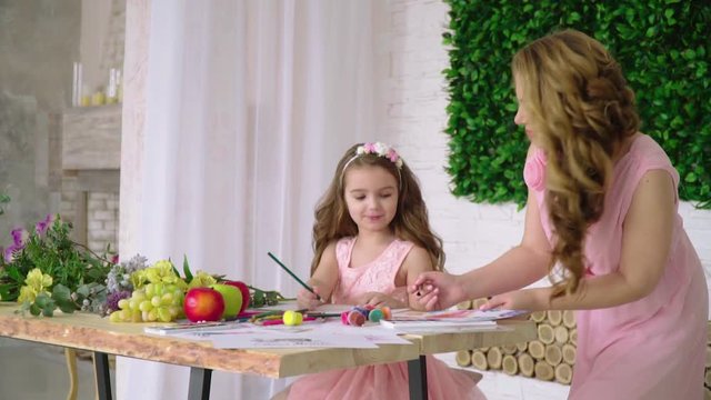 Mom in a pink dress teaches her daughter to paint with brushes