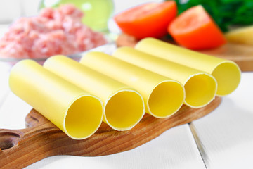 Italian pasta cannelloni. Raw tube for stuffing stuffing surrounded by ingredients for cooking, parmesan cheese, tomato, minced meat on a white wooden table.