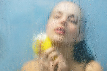 Body care, relief and relaxation concept. Young female model reflects in glass blurred with steam, takes shower, holds sponge for applying gel. Defocused blurry background. Sweat glass surface