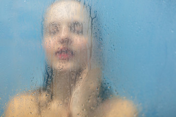 Naked relaxed female stands in shower cabine, takes shower, silhouette through wet glass surface with water drops and stream. Unfocused portrait of woman. Hygiene and freshness concept