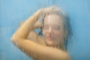Fototapeta premium Young woman refreshes in shower, pamper her skin with hot water, poses in shower cabine with blurred sweat glass. Attractive young female feels relaxation as bathes in bathroom alone