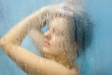 Fototapeta premium Indoor shot of naked female takes shower at bathroom, washes hair, feels relief after hard working day, likes cleannless, takes care of body and skin. Unfocused sillouhette of woman in shower cabine