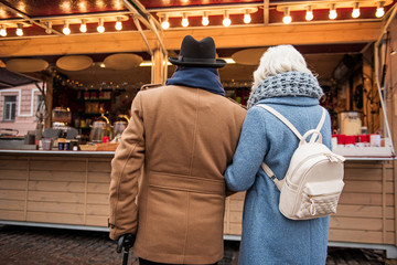 Mature couple is standing near confectionary shop outdoor. Focus on their back
