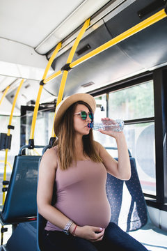 Beautiful young pregnant woman sitting in city bus, drinking water and looking through window. 