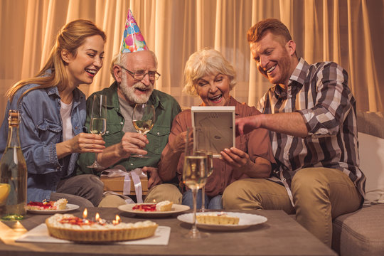 Portrait of happy young woman and man sitting near beaming grandparents. They looking at frame while celebrating birthday. Event concept