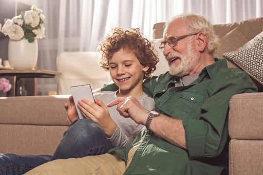 Outgoing kid and cheerful bearded grandfather looking at phone in apartment. Conversation and gadget concept