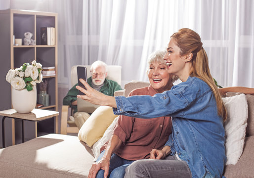 Outgoing grandma and cheerful woman taking selfie by phone while sitting on sofa. Granddad looking at laptop. Entertainment concept