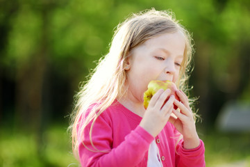 Funny little girl biting an apple outdoors on warm and sunny summer day.