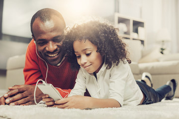 Smiling daddy and his daughter resting on carpet and looking at cellphone with headphones in ears