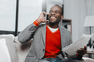 Waist up portrait of pleased guy holding documents while using mobile phone with joy. He is sitting inside