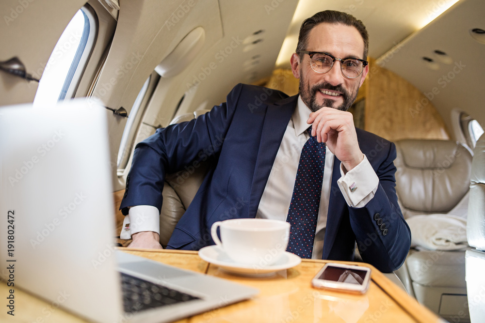 Poster portrait of glad elegant man in airplane sitting at table with cup of coffee and notebook. he is loo