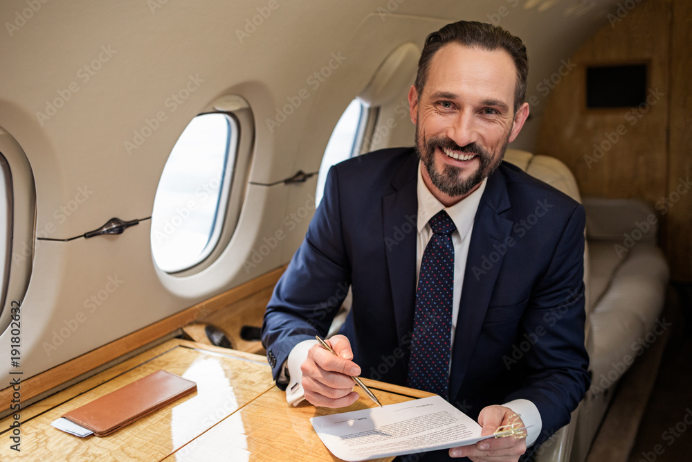 Poster portrait of glad handsome businessman in airplane cabin sitting at table with some documentation. he